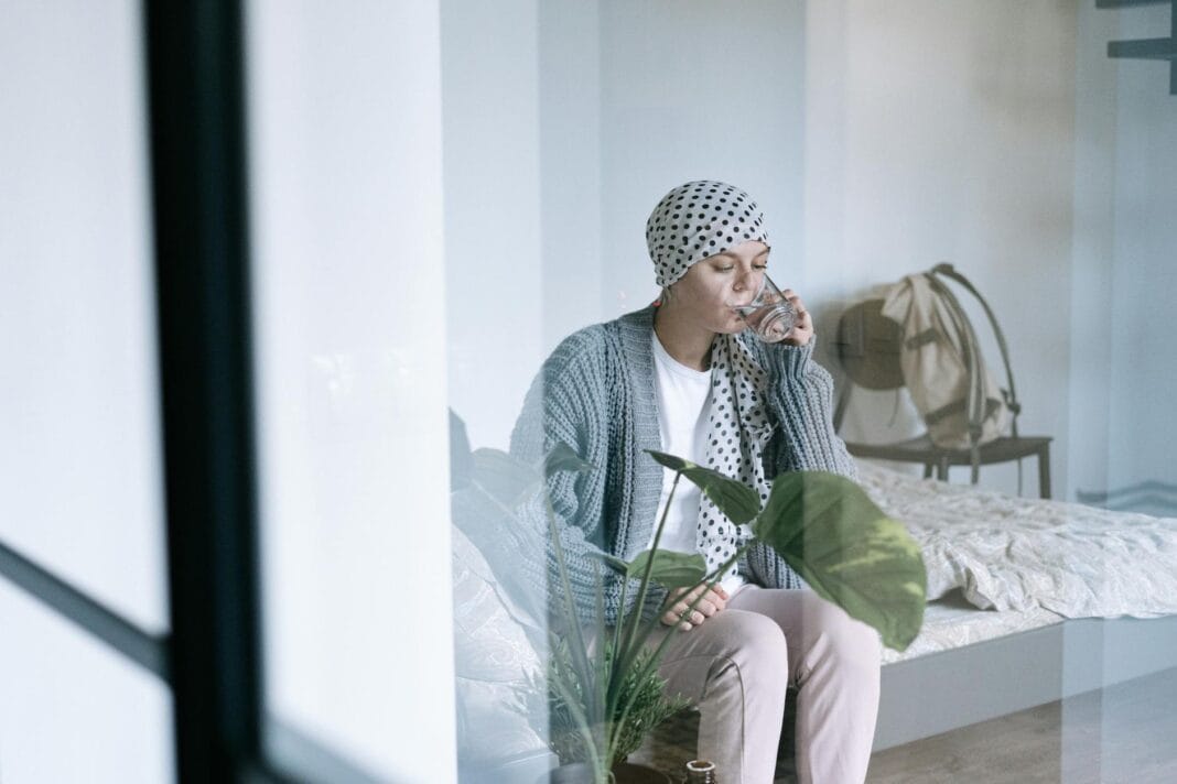 Kobieta w chuście onkologicznej siedzi na łóżku i pije szklankę wody