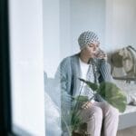 Kobieta w chuście onkologicznej siedzi na łóżku i pije szklankę wody